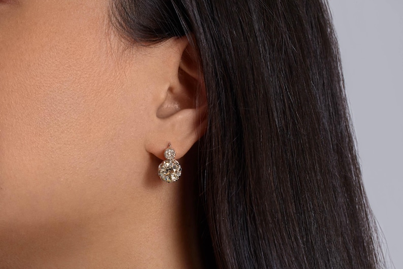 May include: A close-up of a person's ear wearing a pair of diamond drop earrings. The earrings feature a large, round, sparkling diamond at the bottom and a smaller diamond at the top, set in a gold or platinum setting.