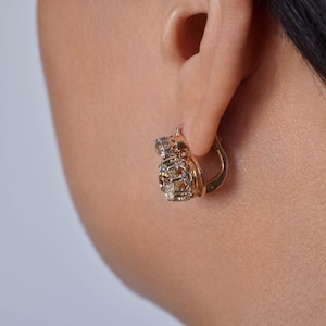 May include: Close-up of a gold-colored hoop earring with a cluster of small, clear and amber-colored stones. The earring is attached to an ear. The background is a light, neutral color.