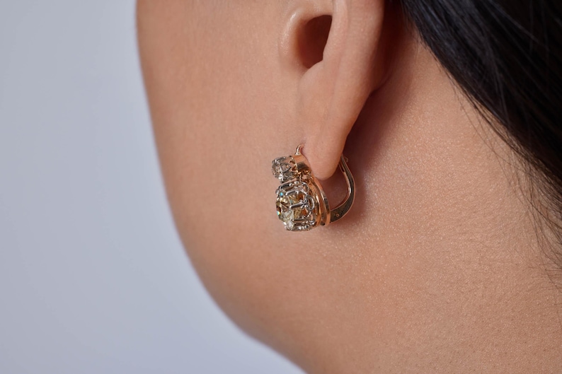May include: Close-up of a gold-colored hoop earring with a cluster of small, clear and amber-colored stones. The earring is attached to an ear. The background is a light, neutral color.