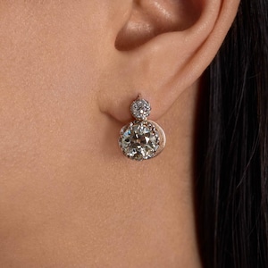 May include: A close-up of a person's ear wearing a diamond earring. The earring has a large, round, clear diamond at the bottom and a smaller diamond at the top. The setting appears to be gold.