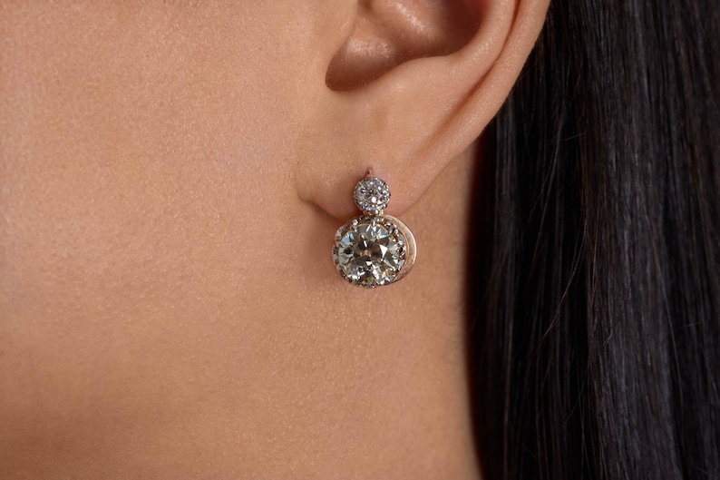 May include: A close-up of a person's ear wearing a diamond earring. The earring has a large, round, clear diamond at the bottom and a smaller diamond at the top. The setting appears to be gold.