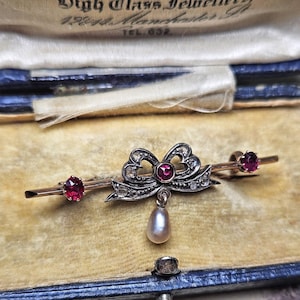 Puede incluir: Broche vintage con diseño de lazo, rubí central y perla colgante. Dos rubíes adicionales a cada lado. El metal tiene tonos plateados y dorados, sobre un fondo de terciopelo.