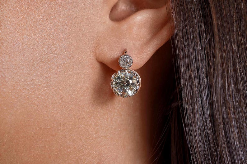 May include: A close-up of a diamond earring worn on an ear. The earring features a large, round, sparkling diamond at the bottom and a smaller diamond at the top. The setting appears to be gold.