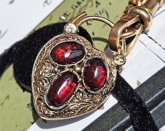 May include: A heart-shaped gold pendant with three oval, deep red gemstones. The pendant has an ornate, engraved design and is attached to a gold chain. A black velvet ribbon is in the background.