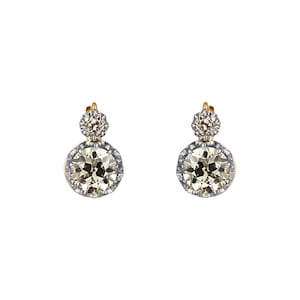 May include: A pair of elegant earrings featuring two round, sparkling gemstones. The earrings have a gold setting with a silver-colored border around the larger, lower stone. The smaller stone sits above the larger one.