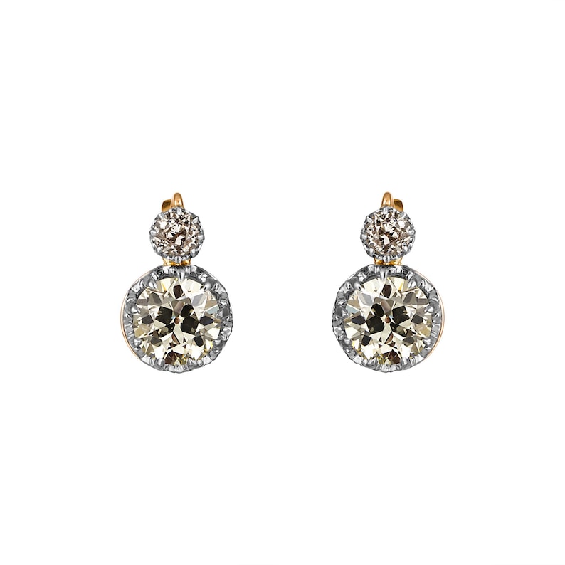 May include: A pair of elegant earrings featuring two round, sparkling gemstones. The earrings have a gold setting with a silver-colored border around the larger, lower stone. The smaller stone sits above the larger one.