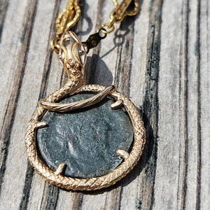 May include: Gold-toned necklace featuring a circular pendant with a dark, textured center. The pendant is encircled by a snake design, with the snake's head at the top. The chain is also gold-toned, creating a cohesive and elegant piece of jewelry.