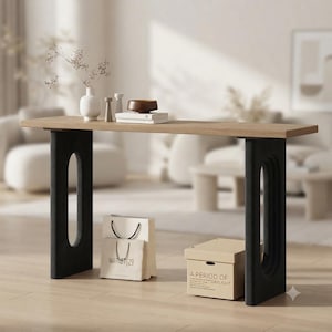 May include: A rectangular console table with a light oak wood top and two black vertical supports featuring large oval cutouts. On the table sits a white vase with dried stems, two small cylindrical objects, and a brown bowl on a white book.