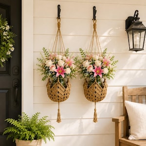 May include: Two hanging planters featuring woven tan jute baskets suspended by knotted cords. Each basket holds a glass vase filled with a floral arrangement of white roses, pink lilies, and green foliage, finished with a decorative bottom tassel.