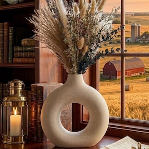 May include: A beige, circular, donut-shaped ceramic vase with a textured finish, holding a tall arrangement of dried grasses, lavender, and fluffy white bunny tails. The vase sits on a wooden surface next to a brass lantern and old books.