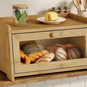 May include: A rectangular bamboo bread box with a clear glass front panel and a small wooden knob. The interior contains various baked goods, including loaves and rolls. The top surface holds a glass jar with leaves and a plate with butter.