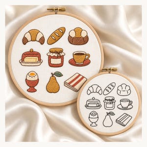 May include: Two circular wooden embroidery hoops featuring breakfast-themed designs on white fabric. The larger hoop displays coloured thread motifs of a croissant, baguette, bun, butter dish, jam jar, coffee cup, egg, pear, and a folded napkin.