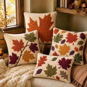 May include: Four decorative throw pillows with cream-colored fabric featuring textured, punch-needle style leaf patterns in shades of orange, yellow, green, and deep purple. The designs include large maple leaves and scattered autumn foliage.