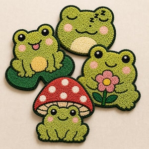 Op de afbeelding: Een verzameling van vier grillige, handgemaakte onderzetters met kikker-thema. De onderzetters hebben verschillende kikkerontwerpen, waaronder een slapende kikker, een kikker met een bloem, een kikker op een waterlelieblad en een kikker onder een paddenstoel. Groen, roze en wit.