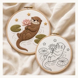 Könnte beinhalten: Zwei runde Stickrahmen mit einem Otter-Motiv. Der größere Rahmen zeigt einen braun gestickten Otter, der eine rosa Lotusblüte hält, umgeben von grünen Seerosenblättern und gelben Sternen. Der kleinere Rahmen zeigt dasselbe Motiv als schwarze Umrisszeichnung.