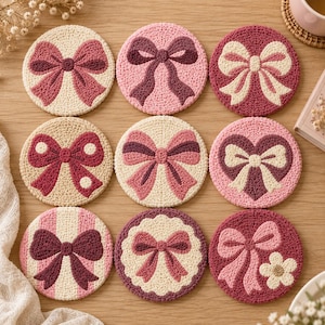Peut inclure: Neuf dessous de verre circulaires au crochet disposés en grille, chacun présentant un motif de nœud unique dans des tons de rose, crème et rouge foncé. Les motifs en laine texturée incluent des rayures, des pois et des blocs de couleur.