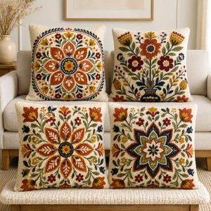 Puede incluir: Cuatro cojines cuadrados con bordado de aguja mágica texturizado sobre una base color hueso. Los diseños incluyen complejos mandalas florales y arreglos botánicos en tonos naranja quemado, amarillo mostaza, azul marino y verde salvia.