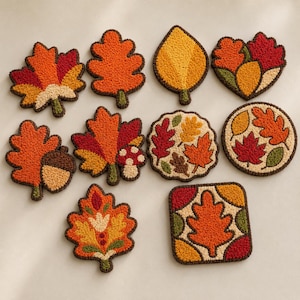 May include: A collection of ten punch needle coasters featuring autumn-themed designs. The set includes various leaf shapes, a round coaster with scattered leaves, and a square coaster, all crafted with textured yarn in shades of orange, red, yellow, and green.