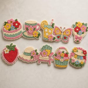 Op de afbeelding: Een set van tien punch needle onderzetters met kleurrijke ontwerpen, waaronder een bloemencake, een theekopje, een glas limonade, een vlinder, een bloemenboeket, een aardbei, een zonnehoed, een stoel, een mandje en een vlaggetjesslinger.