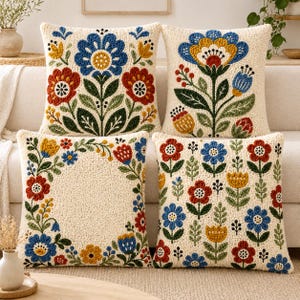 Puede incluir: Cuatro cojines cuadrados de tela texturizada color crema. Cada cojín presenta un patrón floral distinto bordado con hilo azul, rojo, amarillo y verde. Los diseños incluyen grandes flores centrales, arreglos en los bordes y motivos florales repetidos.