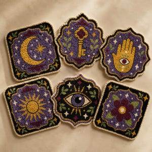 Puede incluir: Un juego de seis posavasos de punch needle con diseños en morado, negro y amarillo. Los motivos incluyen una luna creciente, una llave, una mano de Hamsa con un ojo, un sol, un ojo y una flor, todos bordeados con hilo de color crema.