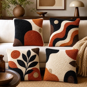 Könnte beinhalten: Vier quadratische Kissen aus strukturiertem, getuftetem Stoff in einer Farbpalette aus Creme, Schwarz, gebranntem Orange und Braun. Die Designs umfassen abstrakte geometrische Formen, wellenförmige Linien und ein zentrales Blattmotiv.