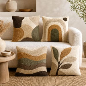 Peut inclure: Un ensemble de quatre coussins carrés en tissu texturé et touffeté dans une palette de crème, beige, vert olive et brun foncé. Les motifs incluent des formes organiques abstraites, un arc-en-ciel, des rayures horizontales ondulées et des feuilles.
