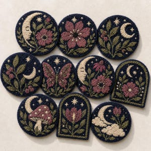 May include: A collection of navy blue, embroidered coasters with various celestial and floral designs. Each coaster features a different motif, including moons, flowers, stars, and a butterfly, in shades of cream, pink, and green.