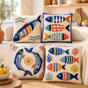 May include: Four square throw pillows featuring cream-colored fabric with embroidered fish designs in blue, red, yellow, and orange. The patterns include single large fish, rows of fish, and fish arranged in circular motifs with coral accents.