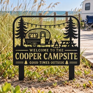 May include: A black metal garden sign featuring a camper, pine trees, a picnic table, and string lights. The text reads WELCOME TO THE COOPER CAMPSITE and GOOD TIMES OUTSIDE, with small pine tree icons flanking the bottom line of text.