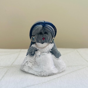 May include: A handmade doll with a gray yarn body, blue hair, and a white lace dress. The doll has small silver earrings and a necklace. The doll's lips are red. The doll is sitting on a white surface.