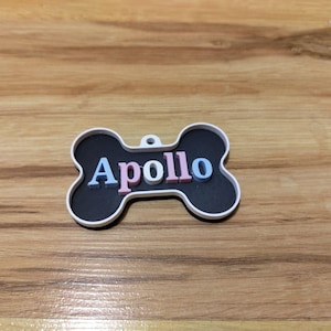 Puede incluir: Una etiqueta para mascotas en forma de hueso con el nombre "Apollo" en letras rosas, blancas y azules. La etiqueta tiene un centro negro y un borde blanco. La etiqueta está sobre una superficie de madera.