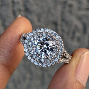 Peut inclure: Une bague argentée avec une grande pierre centrale ronde taille brillant, entourée d'un double halo de pierres plus petites. L'anneau est divisé en deux sections et est incrusté de nombreuses petites pierres étincelantes.