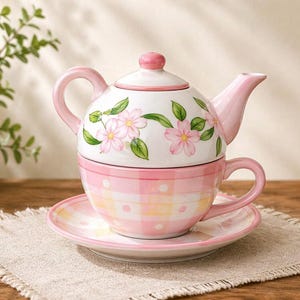 May include: A pink and white floral tea set, including a teapot, teacup, and saucer. The teapot is adorned with pink flowers and green leaves. The teacup and saucer have a pink and white checkered pattern. The set is on a beige linen placemat.