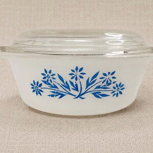 May include: White Pyrex bowl with a clear glass lid. The bowl features a blue floral design. The bowl has two handles and is sitting on a neutral-colored surface. A vintage kitchenware item.