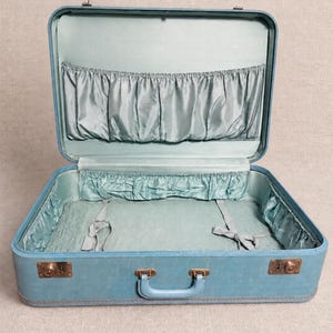 May include: An open, light blue suitcase with a matching interior lining. The interior features gathered fabric pockets along the lid and base, along with two fabric tie-down straps at the bottom. The exterior includes a central handle and metal latches.