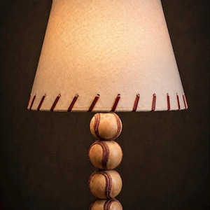 May include: A baseball-themed table lamp with a beige fabric shade and a base made of stacked baseballs. The shade is trimmed with red stitching, and the lamp sits on a wooden surface.