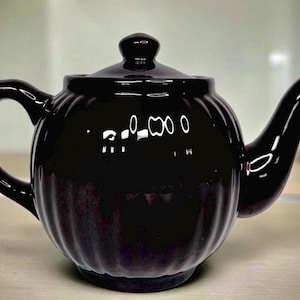 May include: A glossy black ceramic teapot with a lid and a curved spout. The teapot has a rounded body with vertical ridges and a handle on the side. The teapot is designed for brewing tea.