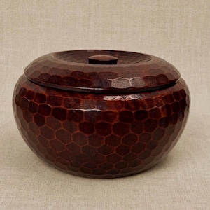 May include: A round, dark brown wooden container with a lid. The container and lid have a textured, honeycomb-like pattern. The lid has a small, round knob in the center. The container is sitting on a neutral-colored surface.