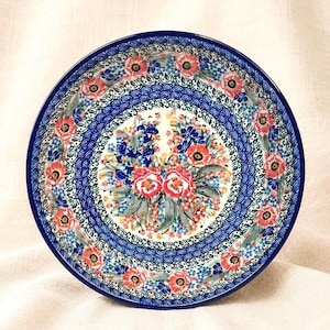 May include: A round ceramic plate with a vibrant floral design. The plate features a deep blue rim and a central pattern of red, blue, and white flowers. The intricate design includes detailed patterns and a white background.