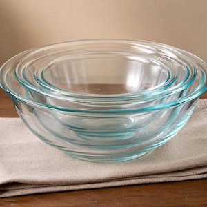 May include: A set of four clear glass mixing bowls nested together on a beige linen napkin. The bowls are different sizes, with a rounded shape and a slight blue tint around the rims. The bowls are sitting on a wooden surface.
