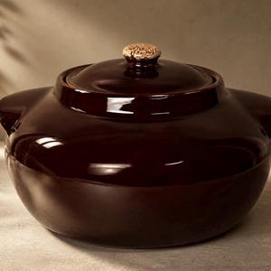 May include: A dark brown ceramic cooking pot with a lid. The pot has two handles and a textured knob on the lid. The pot is round and has a glossy finish.