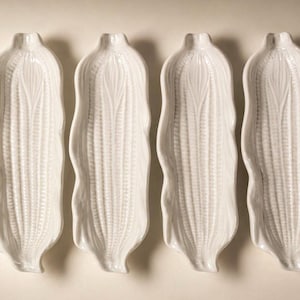 May include: Four white ceramic corn-shaped serving dishes. Each dish has a detailed embossed design mimicking the texture of corn. The dishes are arranged side by side on a light-colored surface, ideal for serving food.