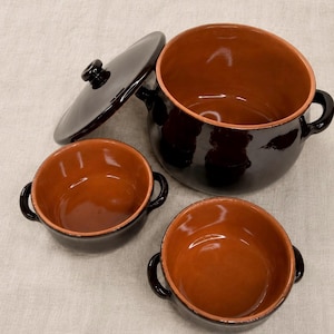 May include: A set of brown ceramic cookware, including a large pot with a lid and two smaller bowls. The interior of the cookware is a lighter brown, contrasting with the dark brown exterior. The pot has two handles.