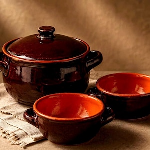 May include: A set of three brown ceramic cookware pieces. The set includes a lidded pot and two bowls with handles. The interiors of the bowls are a vibrant orange. The pieces are arranged on a neutral-colored cloth.