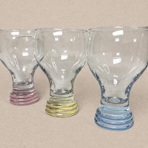 May include: Three clear glass goblets with a bulbous bowl and a stem with colorful horizontal stripes. The stems are pink, yellow, and blue. The goblets are likely for beverages.