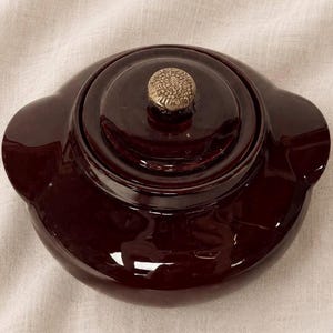 May include: A dark burgundy ceramic casserole dish with a lid and a decorative gold-colored knob. The dish has a glossy finish and two side handles. The lid fits snugly on top of the dish.