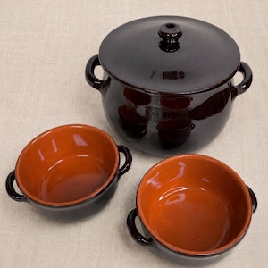 May include: A set of three ceramic cookware pieces. The set includes a large, dark brown pot with a lid and two smaller bowls. The interiors of the bowls are a warm orange color, contrasting with the dark exterior. The pot and bowls have handles.