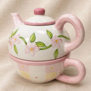 May include: A white ceramic teapot and teacup set with pink accents. The teapot features a lid with a pink knob and is decorated with pink floral and green leaf designs. The teacup has a pink handle and a checkered pattern.