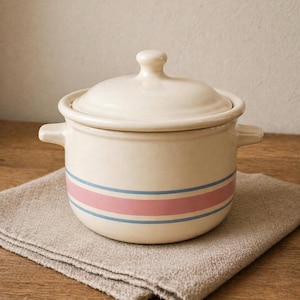 May include: Cream-colored ceramic pot with a lid and two handles. The pot features a pink stripe between two blue stripes around the middle. The pot is sitting on a folded, tan linen cloth. A classic kitchenware item.